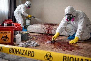 Deux techniciens en combinaison de protection nettoient et désinfectent une chambre après un décès, avec matériel de décontamination et signalisation biohazard.