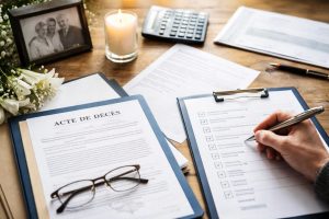 Main remplissant une checklist de démarches après décès avec un acte de décès sur un bureau, une bougie allumée, des fleurs blanches et une photo de famille en arrière-plan