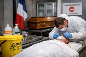 Thanatopracteur en tenue de protection réalisant un soin de présentation sur un défunt en chambre funéraire, avec signalétique d’accès interdit et matériel de sécurité biologique.