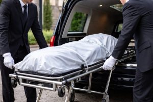 Deux professionnels des pompes funèbres en tenue sombre et gants blancs chargent un corps recouvert d’un linceul sur un brancard dans un véhicule funéraire, en extérieur.