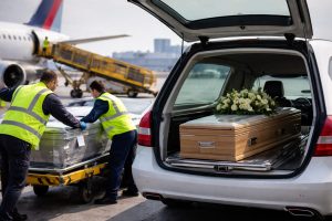Agents en gilet fluorescent transférant un cercueil sur un chariot près d’un avion, à côté d’un véhicule funéraire, lors d’un rapatriement de corps.