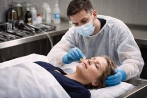 Thanatopracteur portant des gants et un masque réalisant un soin de conservation et une préparation esthétique du visage d’une défunte en environnement médicalisé.